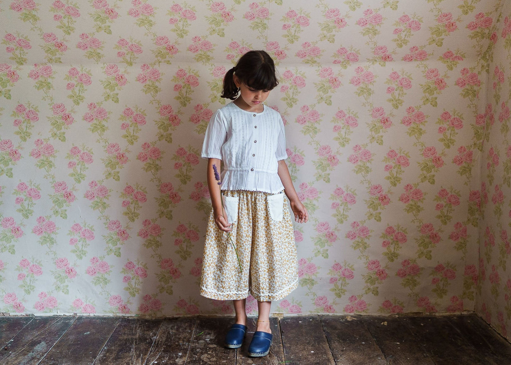Young girl wearing an off-white blouse made from organic cotton paired with a vintage-style floral skirt, standing in a softly lit room with floral wallpaper.