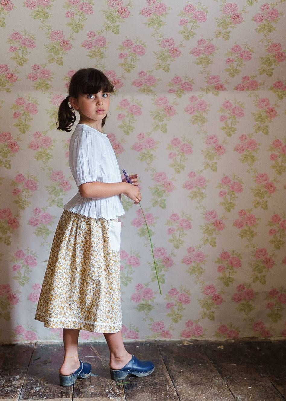 Young girl in vintage style natural cotton clothing stands on rustic wooden floor against floral wallpaper, holding a wildflower, reflecting slow, ethical children's fashion.
