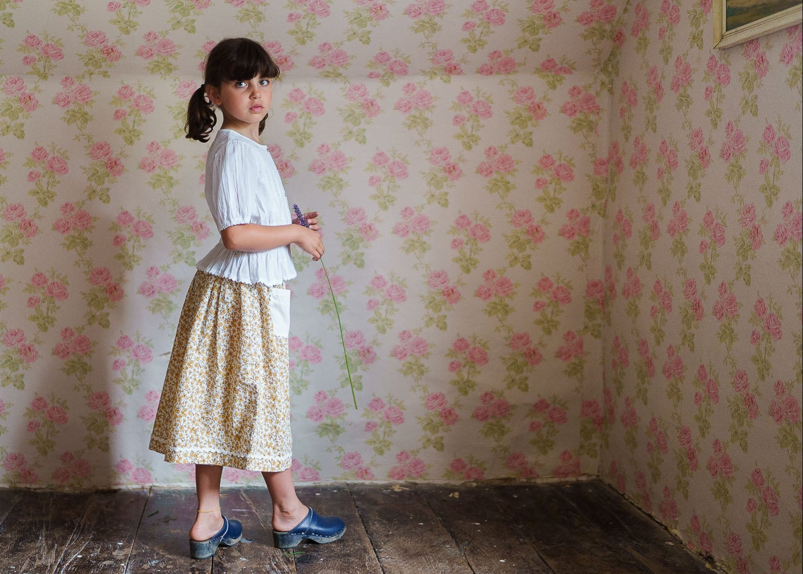 Young girl wearing an off-white organic cotton blouse paired with a vintage-style floral skirt, standing barefoot on a rustic wooden floor against a floral wallpaper backdrop.