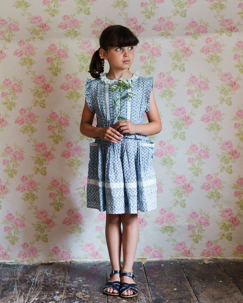 Young girl in a blue dress standing against a floral wallpapered wall.