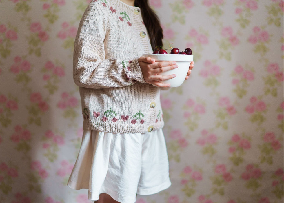 Child wearing a vintage-style hand-knitted cotton cardigan with cherry embroidery, holding a bowl of cherries, showcasing sustainable and organic children's clothing.