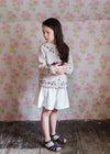 Young girl in vintage style organic cotton clothing wearing a hand-knitted cardigan with cherry details, standing against a floral wallpaper background.