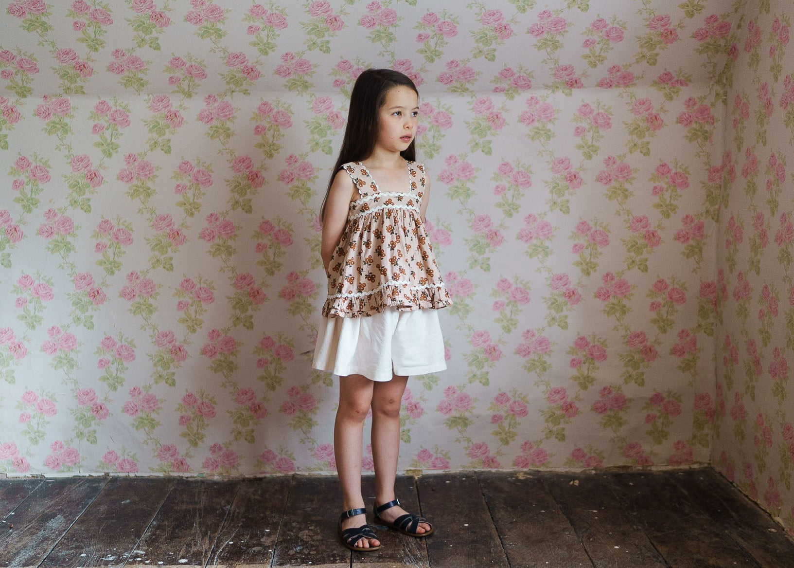 Young girl wearing a vintage-style floral organic cotton sun top paired with white shorts, standing in a softly lit room with pink floral wallpaper and wooden floorboards.