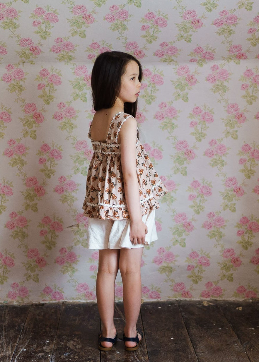 Young girl wearing a vintage-style floral organic cotton sun top with white shorts, standing against a pink and green floral wallpaper in a natural, ethical children's clothing setting.