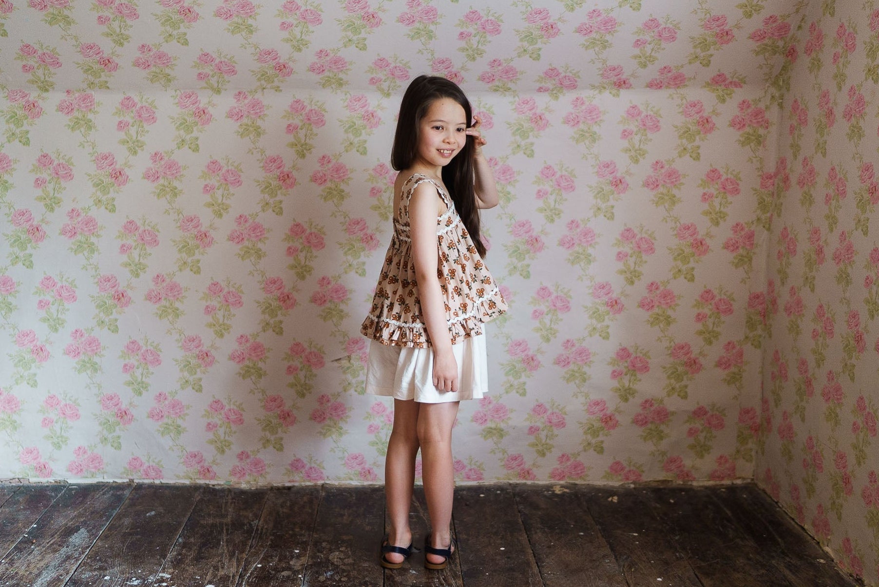 Young girl wearing a vintage-style floral organic cotton top and white skirt, standing barefoot on rustic wooden floor against a pink floral wallpaper backdrop.