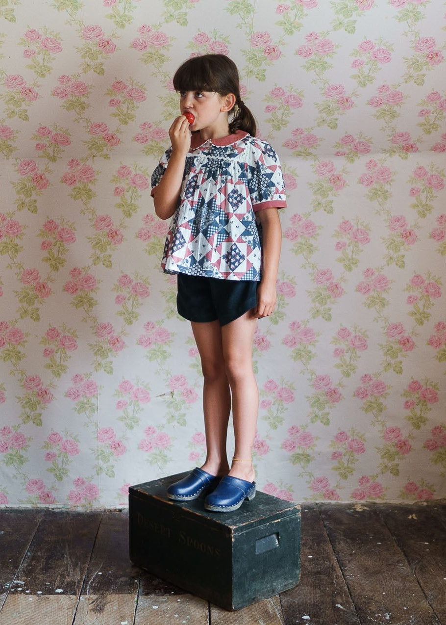 Young girl wearing a vintage style patchwork floral blouse made from organic cotton, standing on a wooden box against a floral wallpaper, showcasing sustainable children's clothing.