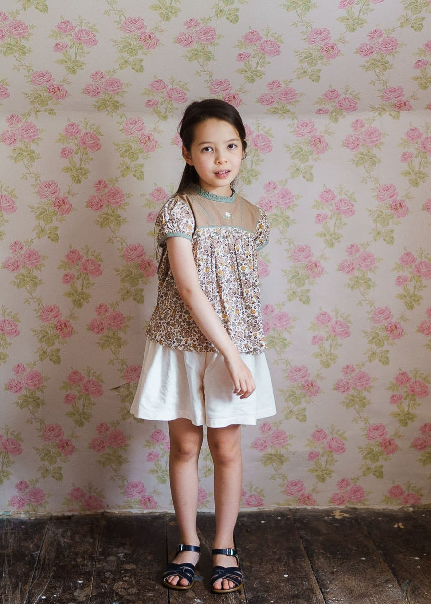 Young girl wearing a vintage-style floral blouse made from organic cotton, paired with a white skirt, standing against a floral wallpaper background in a natural, ethical children's clothing setting.