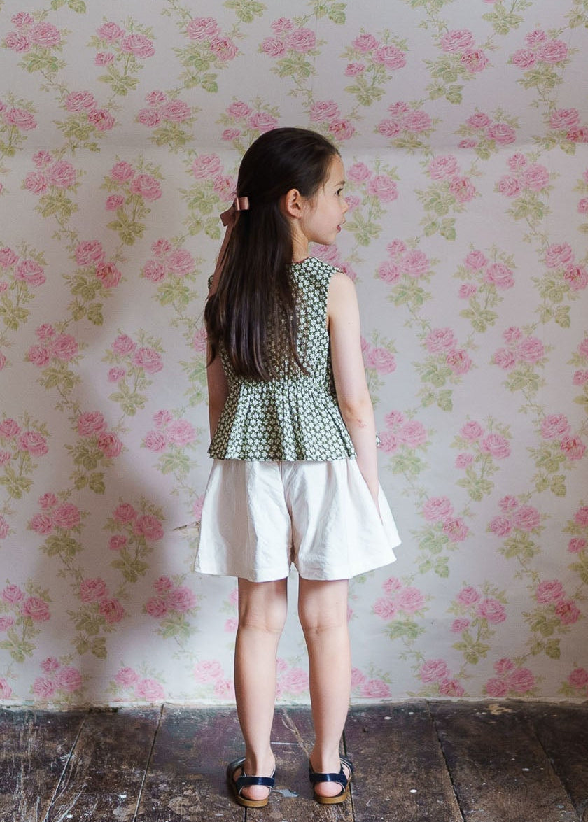 Young girl wearing vintage style organic cotton cream culottes paired with a patterned green top, standing against a floral wallpaper background in a natural, ethical children’s clothing setting.