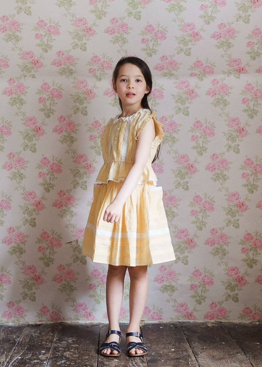 Young girl wearing a vintage style yellow organic cotton blouse and skirt set, standing against a floral wallpaper background, showcasing sustainable children’s clothing.