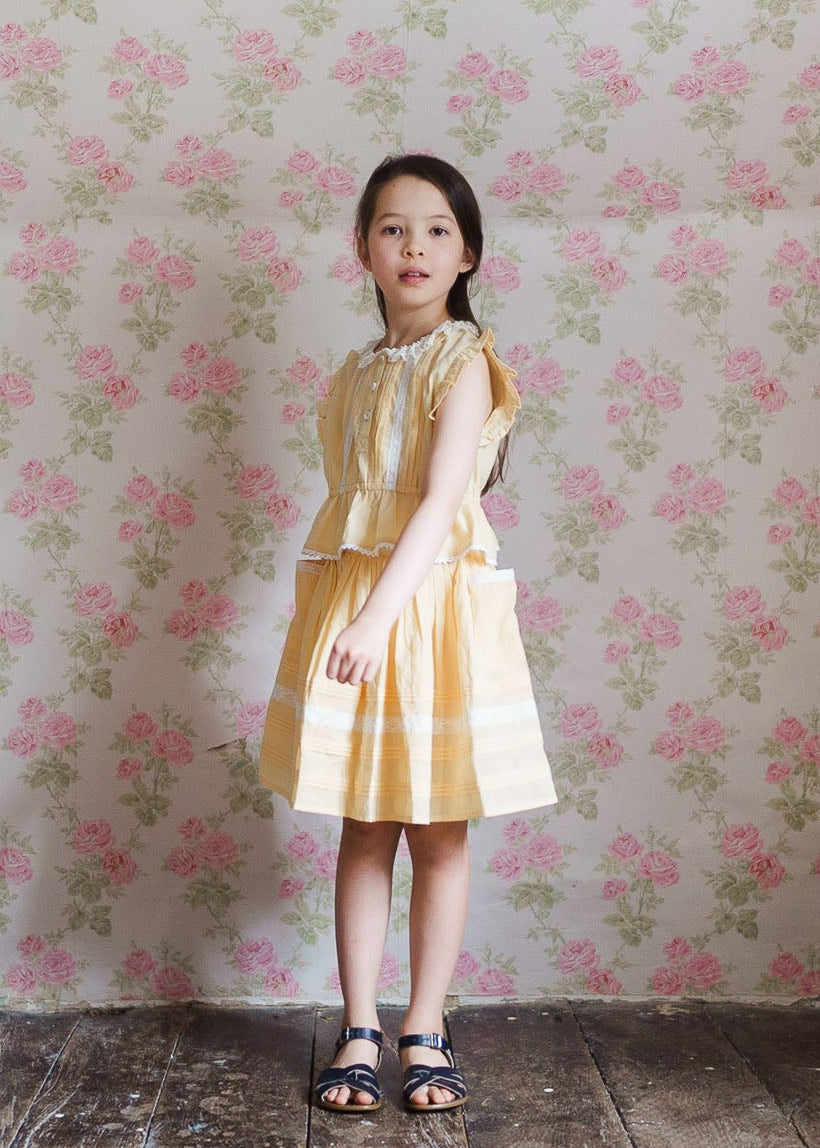 Young girl wearing a vintage style yellow organic cotton skirt and matching top standing against a floral wallpaper, showcasing natural and sustainable children's clothing.