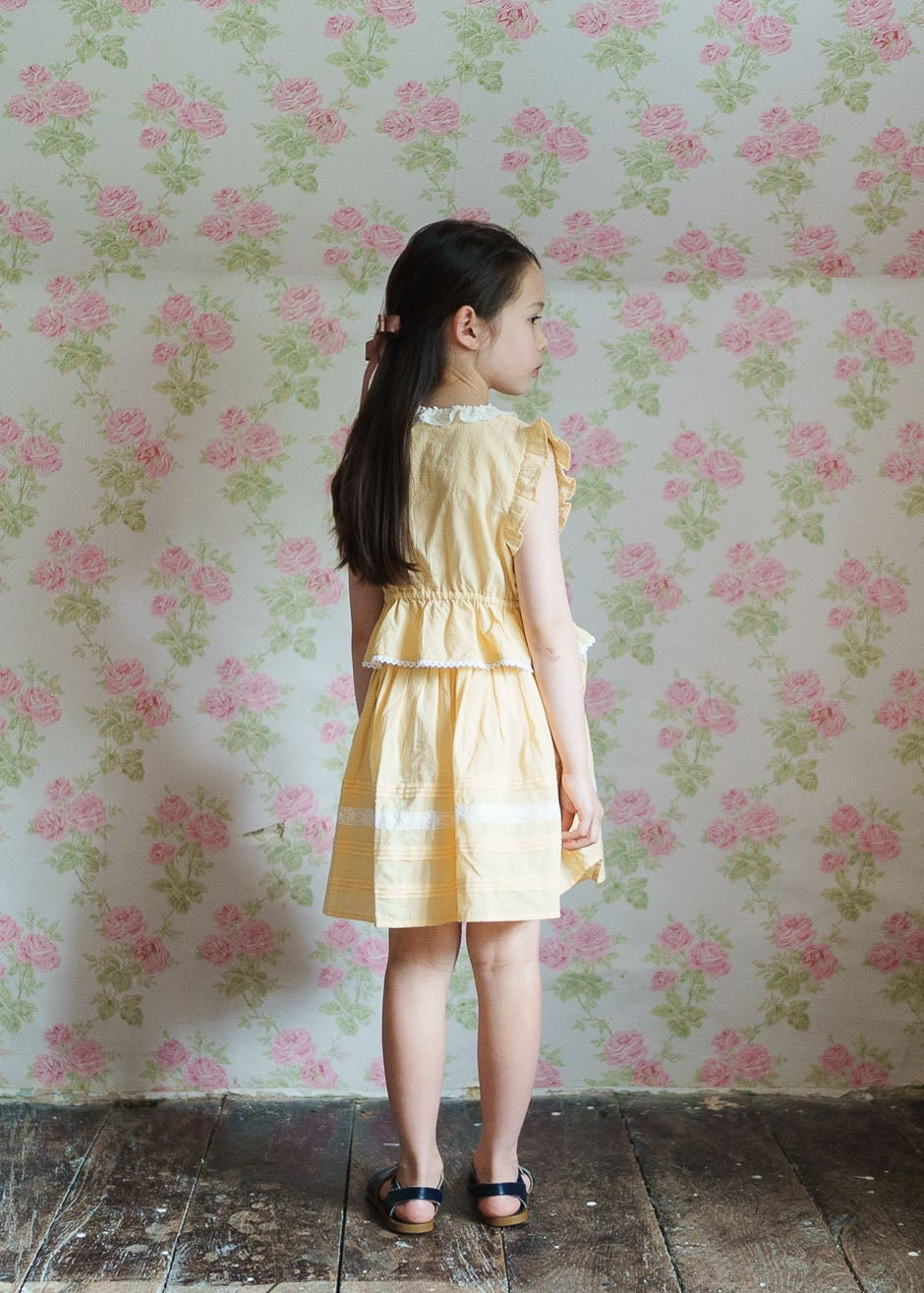 Young girl wearing a vintage style butter yellow organic cotton blouse and skirt set, standing on rustic wooden floor against floral wallpaper, showcasing natural children's clothing.