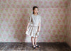 Young girl wearing a vintage-inspired, natural cotton blouse with straw check pattern paired with a matching skirt, standing against floral wallpaper in a rustic room.