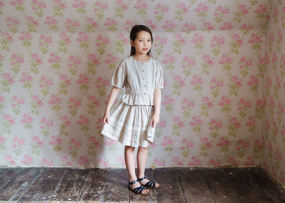 Young girl wearing a vintage-inspired, natural cotton blouse with straw check pattern paired with a matching skirt, standing against floral wallpaper in a rustic room.