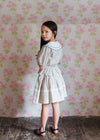 Young girl wearing a vintage-inspired straw check blouse with a white collar, standing in a softly lit room with floral wallpaper, showcasing natural, organic children’s clothing.