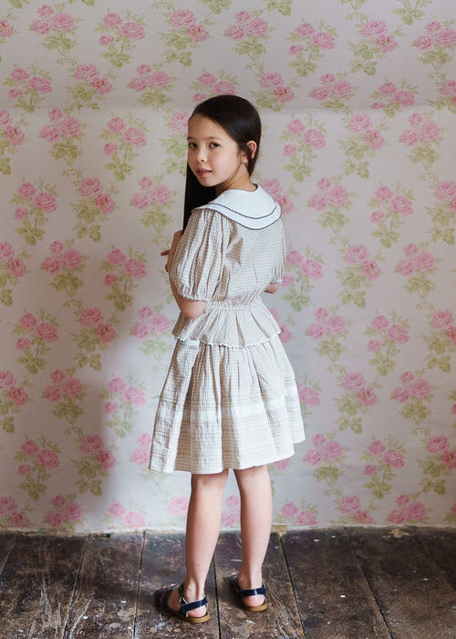 Young girl wearing a vintage-inspired straw check blouse with a white collar, standing in a softly lit room with floral wallpaper, showcasing natural, organic children’s clothing.