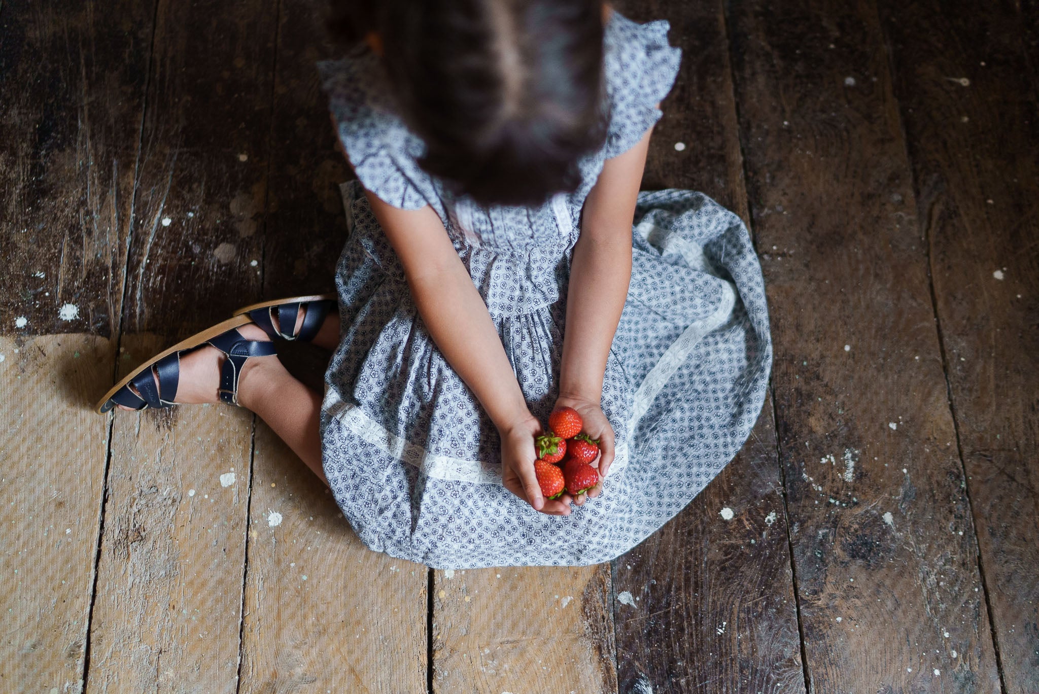 Young girl wearing a vintage style blue floral organic cotton skirt holding fresh strawberries while sitting on a rustic wooden floor, embodying natural and ethical children's clothing.
