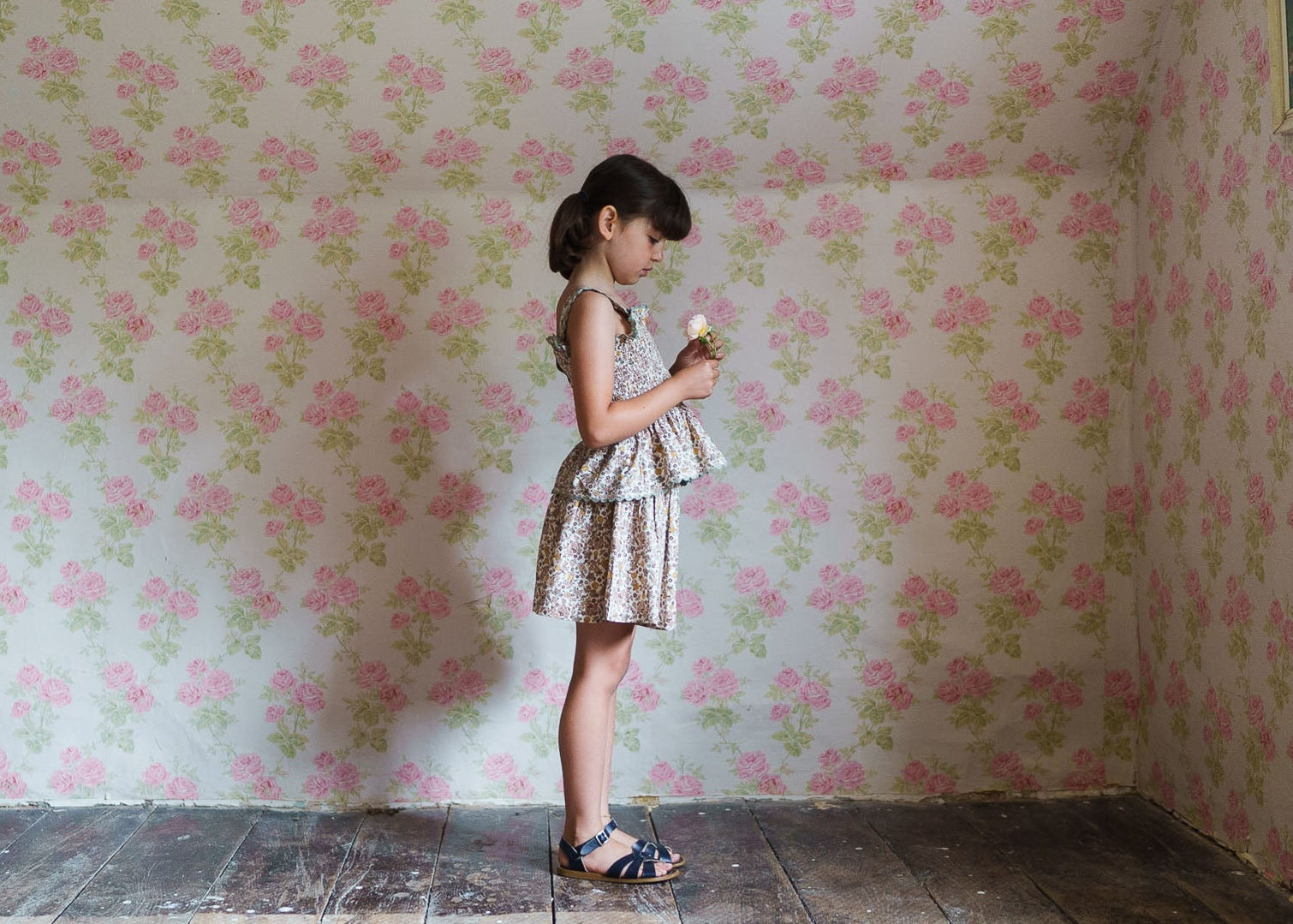 Young girl wearing floral organic cotton culottes and top in a vintage-style room with coordinating floral wallpaper, showcasing slow, natural, and ethical children's clothing.