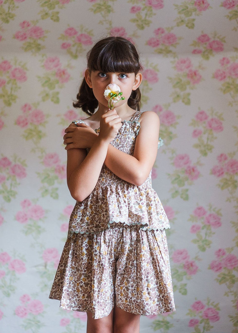 Young girl wearing organic cotton floral culottes and top in vintage style children’s clothing, holding a flower against a soft floral wallpaper background.