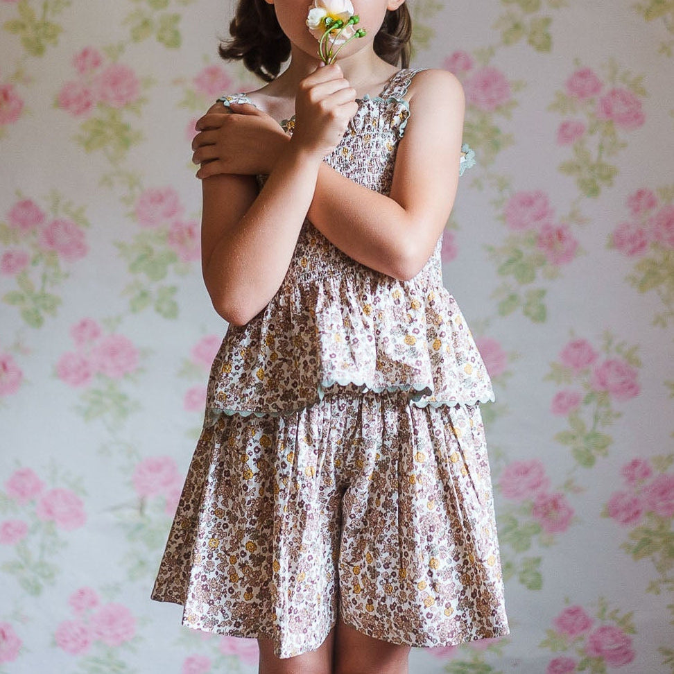 Young girl in a vintage style organic cotton floral top and skirt set, holding a white flower against a soft floral patterned background, evoking natural childhood innocence.