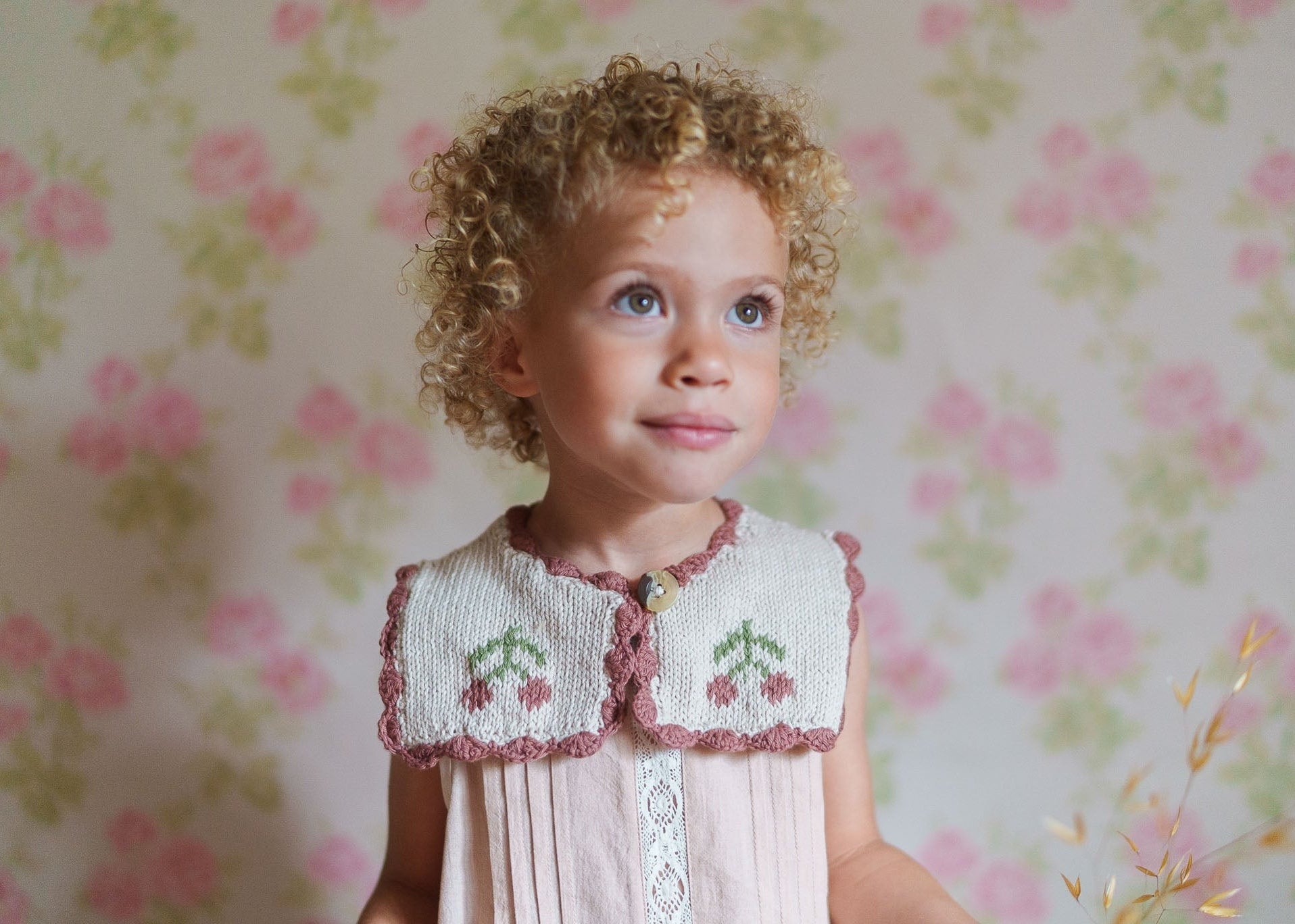 Young child with curly hair wearing a vintage-style hand-knitted cotton sailor collar over a soft pink dress, embodying organic and sustainable children's clothing.