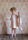 Young child in a vintage-style organic cotton dress holding a hand-knitted scarf, standing against floral wallpaper in a cozy, natural setting.