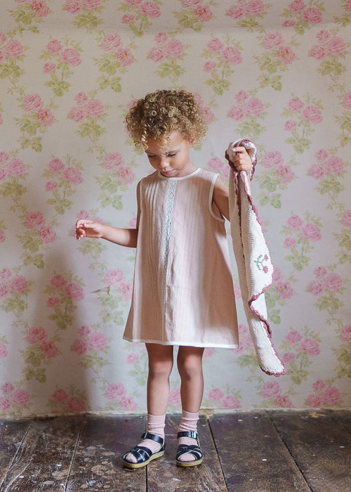 Young child in a vintage-style organic cotton dress holding a hand-knitted scarf, standing against floral wallpaper in a cozy, natural setting.