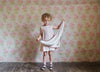 Young child wearing vintage style organic cotton dress holding a soft cherry-colored hand-knitted scarf in a room with floral wallpaper, showcasing sustainable children's clothing.