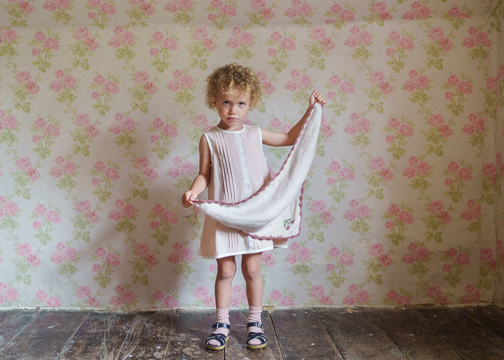 Young child wearing vintage style organic cotton dress holding a soft cherry-colored hand-knitted scarf in a room with floral wallpaper, showcasing sustainable children's clothing.
