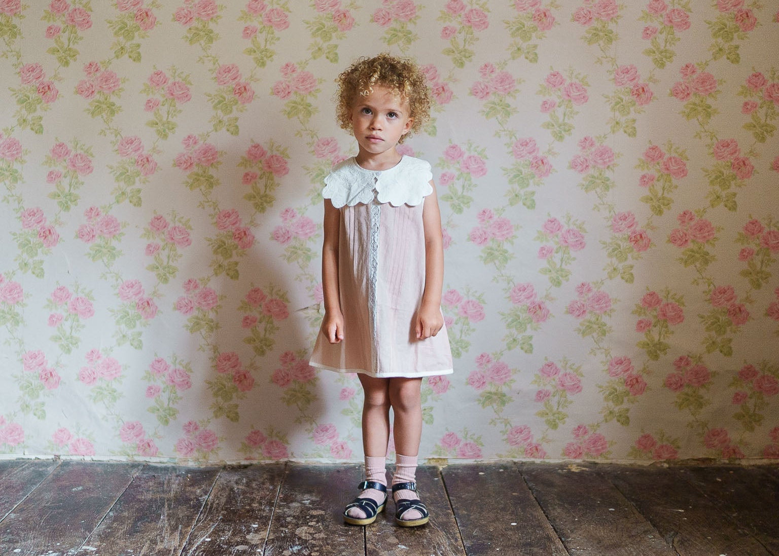 Young girl wearing a vintage style girls dress with an embroidered scalloped cotton collar, standing against a floral wallpaper background on a wooden floor.