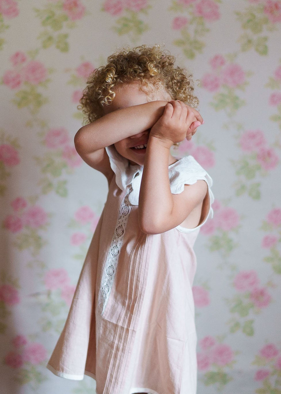 A young child wearing a vintage style dress made from organic cotton with an embroidered scalloped collar, standing against a soft floral wallpaper background.