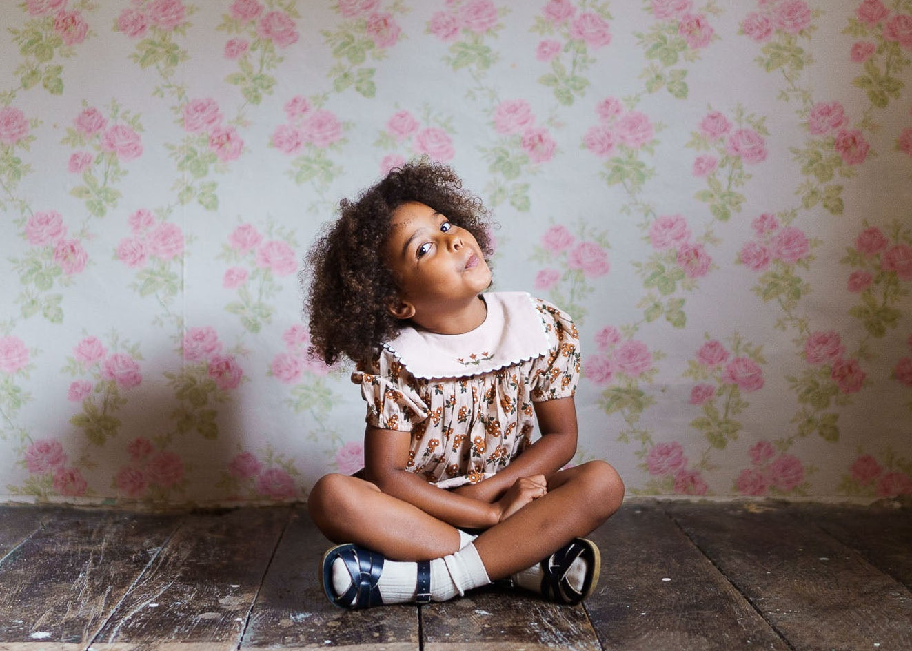 Young girl sitting cross-legged on wooden floor wearing a vintage style organic cotton romper with floral pattern, against a soft pink floral wallpaper background.