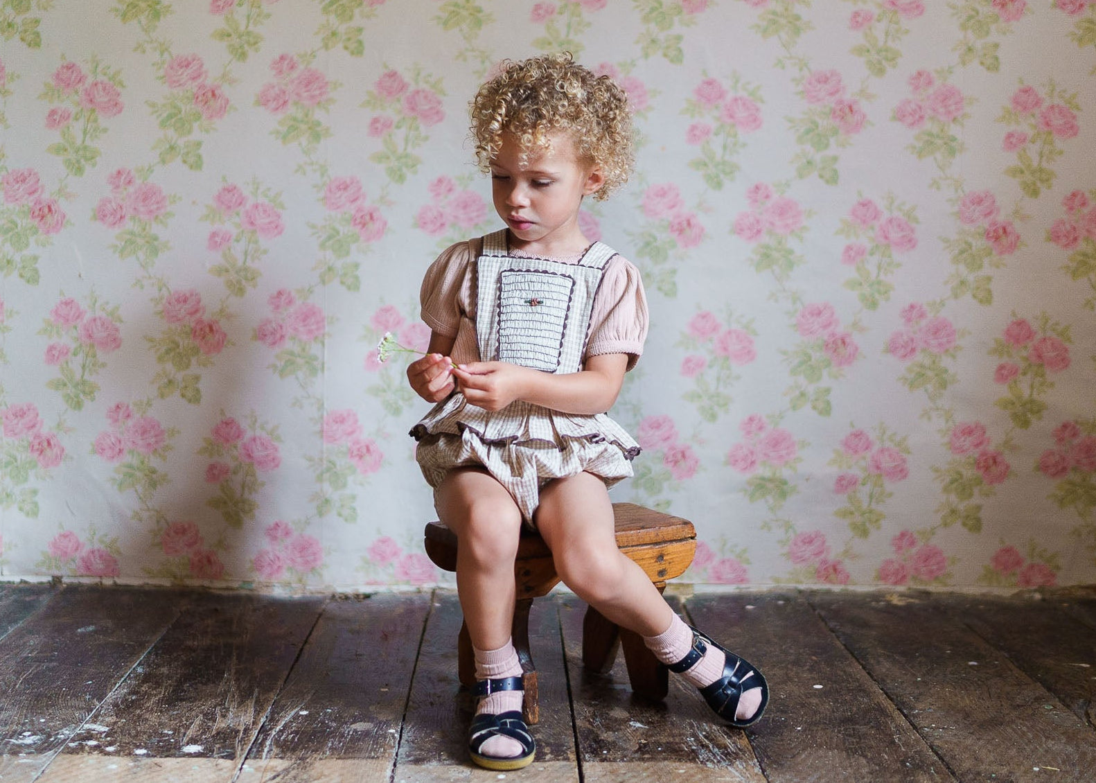A young child with curly hair sitting on a wooden stool wearing a vintage-style straw check romper made from organic cotton, against a floral wallpaper background.