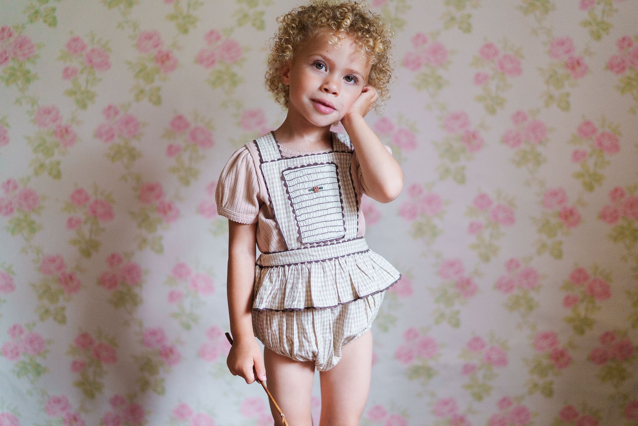 Young child wearing vintage style organic cotton clothing with delicate pink and white patterns, standing against a floral wallpaper background in a softly lit room.