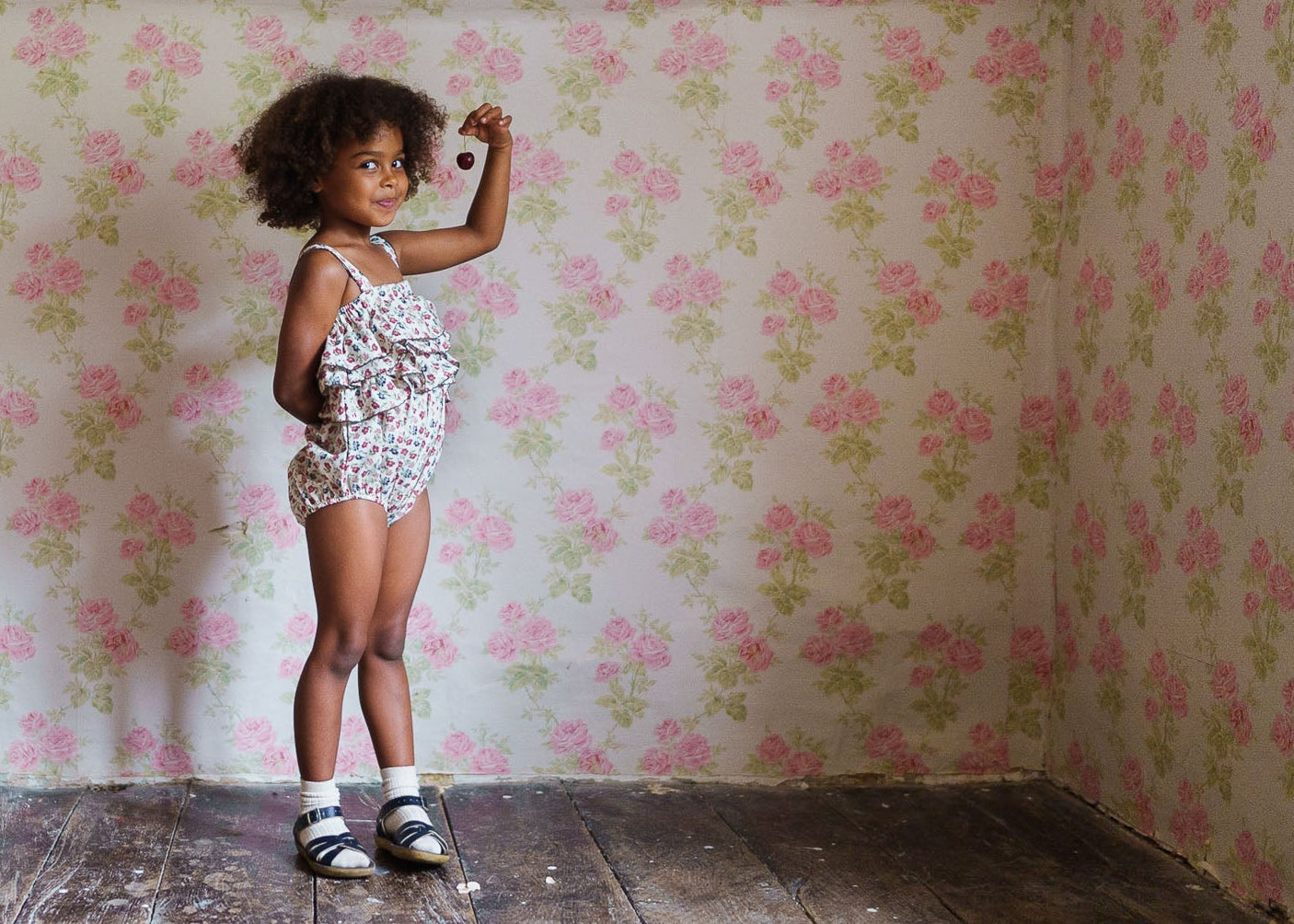 Young girl wearing a floral organic cotton romper in a vintage-style room with floral wallpaper, showcasing sustainable and ethically made children’s clothing.