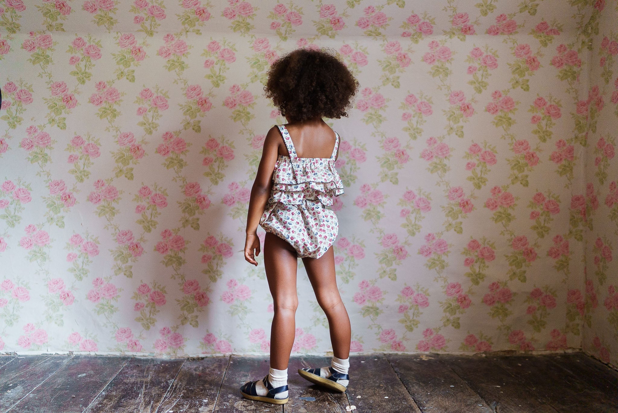 Young girl wearing a floral organic cotton romper with ruffle details, standing in a vintage style room with floral wallpaper, showcasing sustainable, ethically made children's clothing.