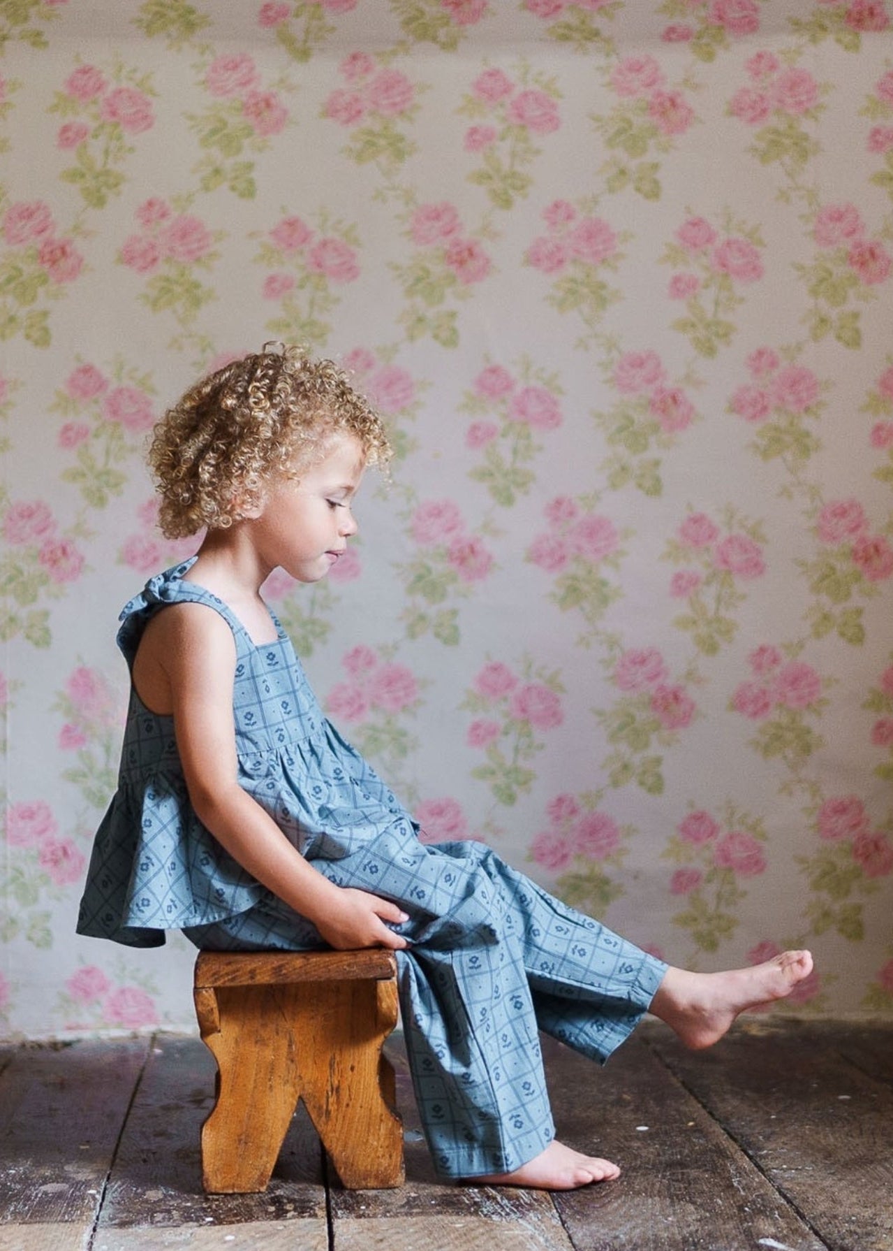A young child with curly hair wearing organic cotton blue patterned trousers and top, sitting barefoot on a rustic wooden stool against a vintage floral wallpaper backdrop.