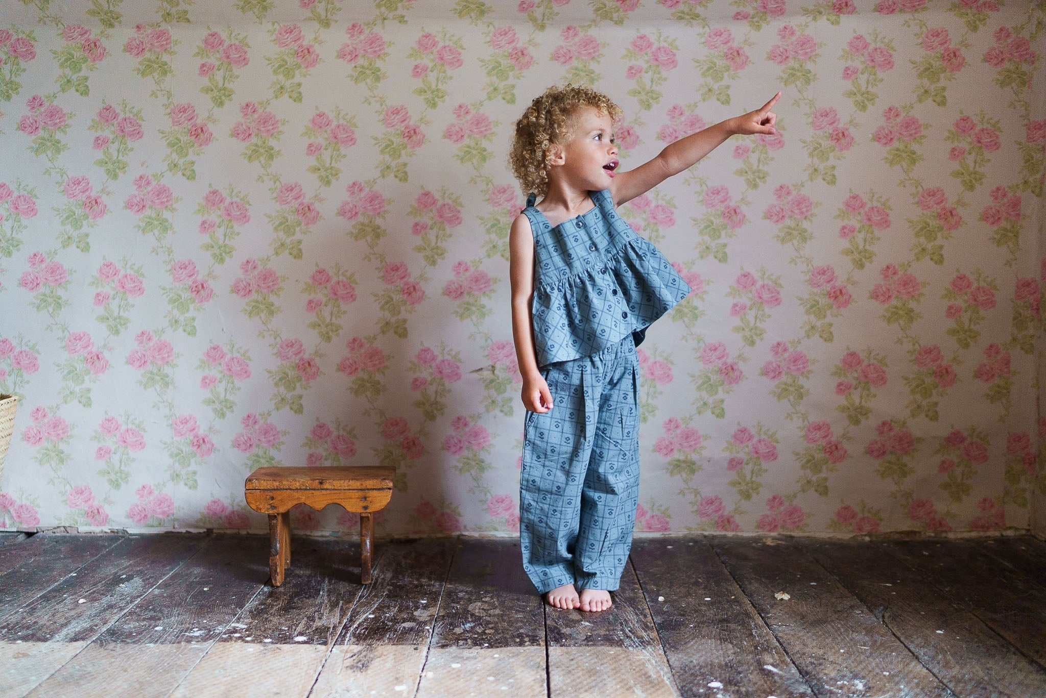 Child wearing a vintage style organic cotton floral top and matching pants, standing barefoot on worn wooden floor with pink floral wallpaper background, pointing ahead.