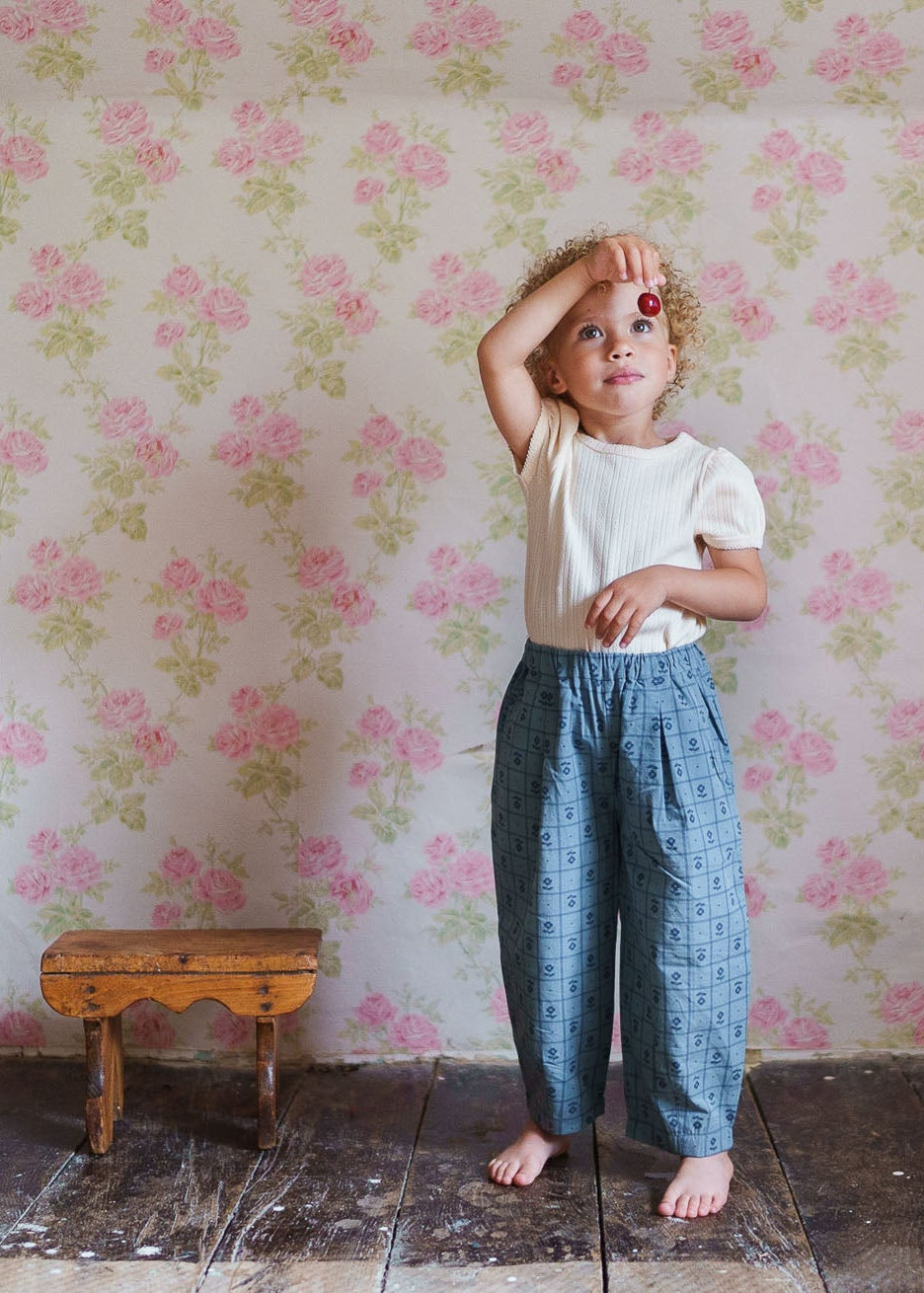 A young child wearing loose, vintage-style organic cotton trousers with a subtle floral pattern, standing barefoot on rustic wooden floor against a soft pink floral wallpaper.