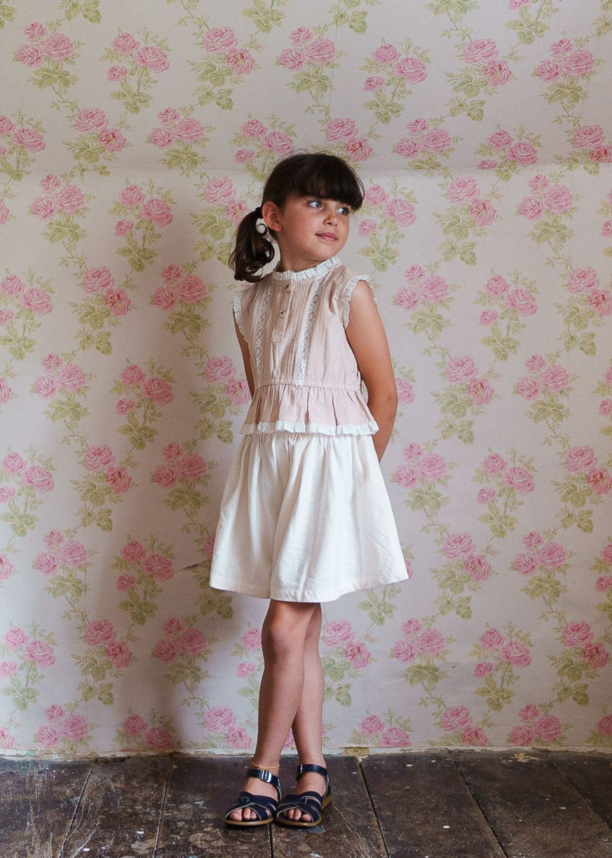Young girl wearing a vintage-style cream organic cotton culottes outfit standing against a floral wallpaper, showcasing sustainable children's clothing with an elegant and timeless look