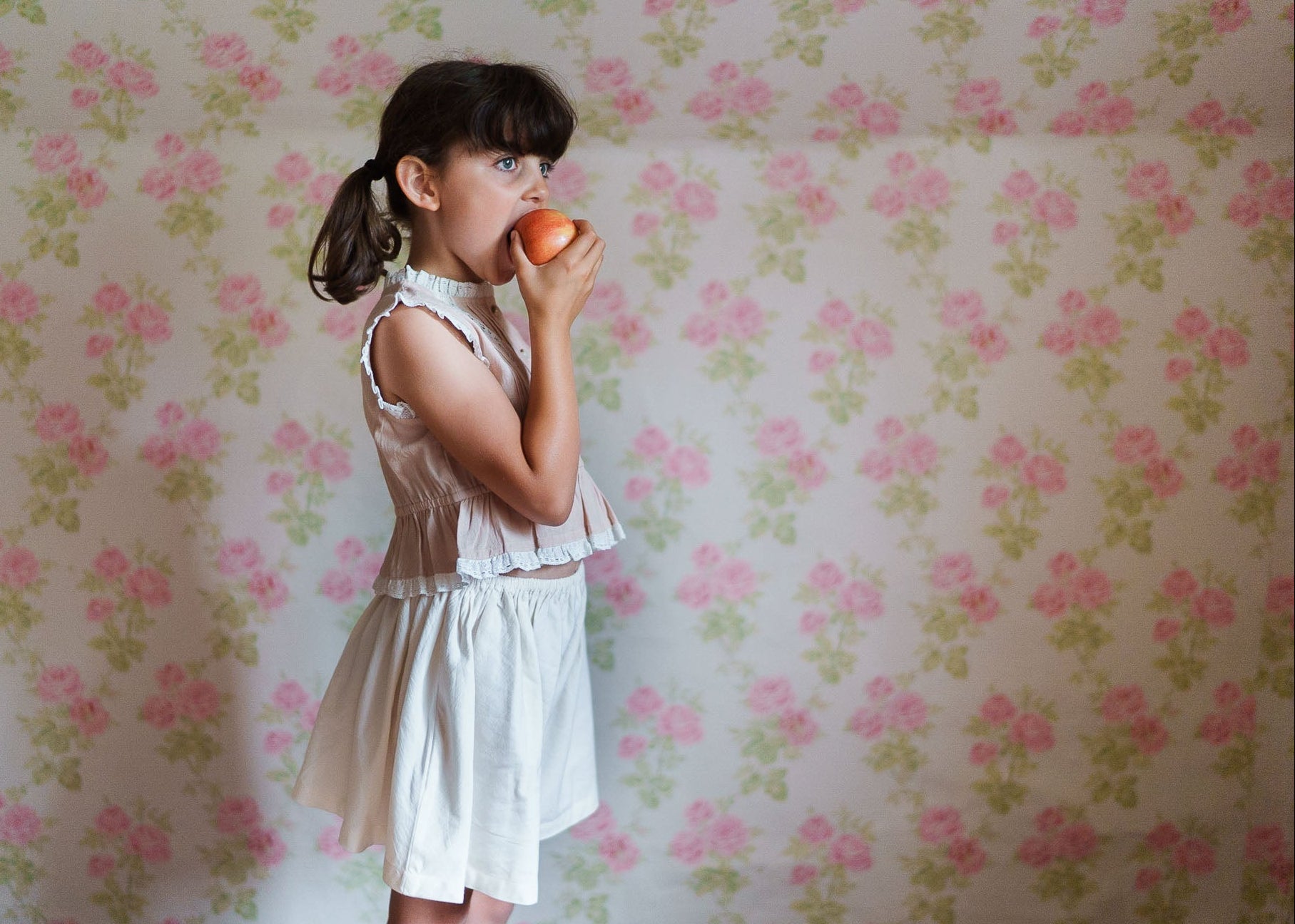 Young girl wearing cream organic cotton culottes and vintage style top, standing against floral wallpaper while holding an apple in a natural, ethical children's clothing setting.