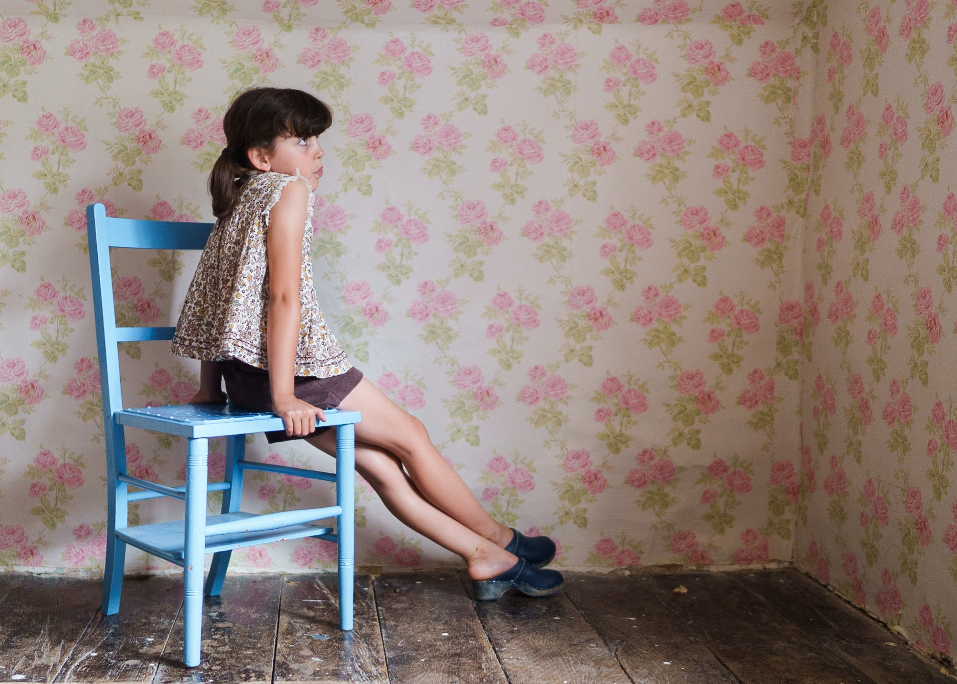 A young girl wearing a vintage style floral blouse made from organic cotton sits on a light blue wooden chair against a softly patterned floral wallpaper in a cozy, natural setting.