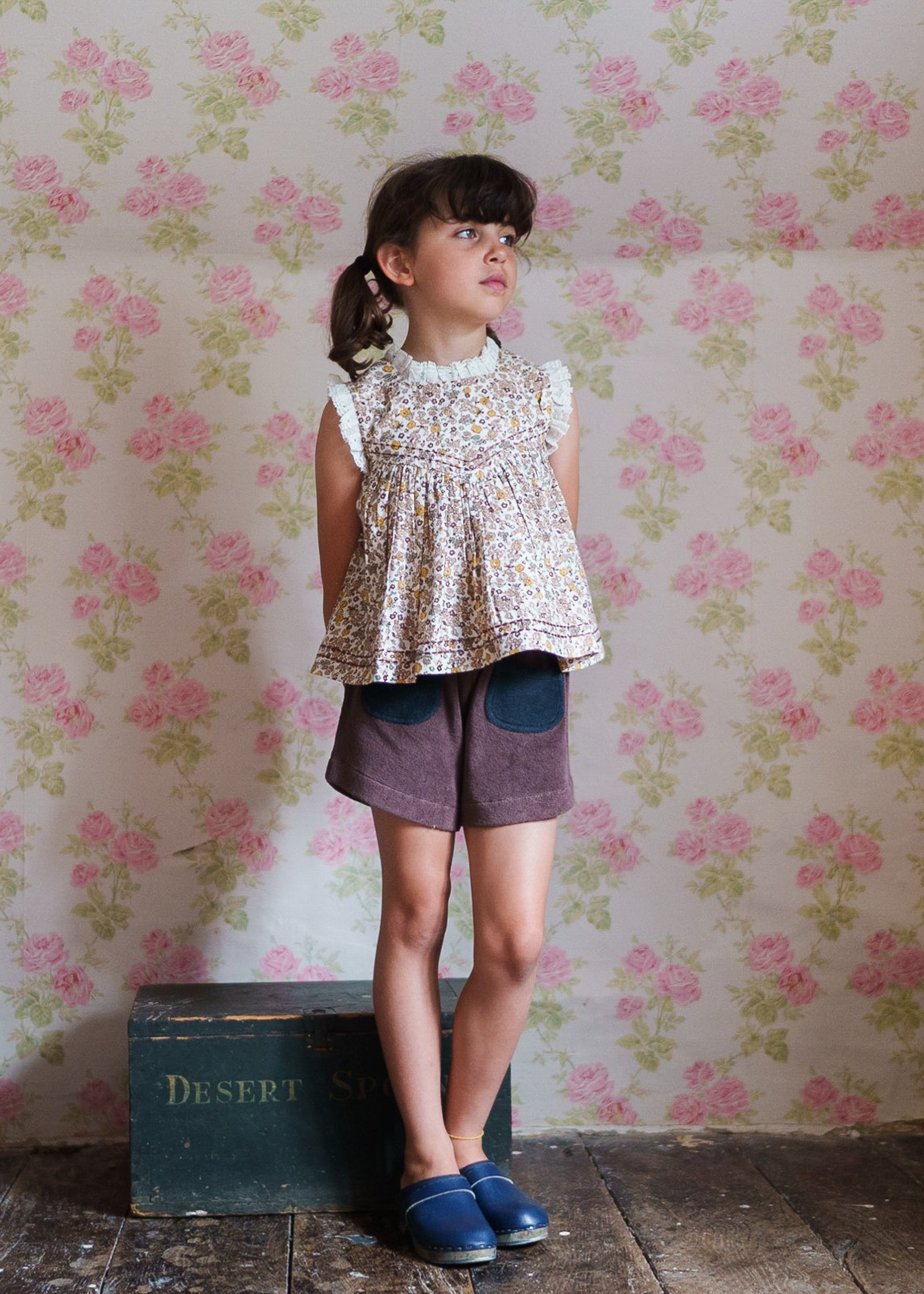 Young girl wearing a vintage style floral organic cotton blouse paired with brown shorts, standing on rustic wooden floor against a rose patterned wall, showcasing sustainable children's clothing.