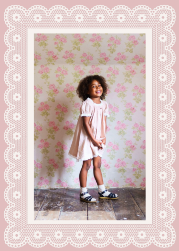 Young girl in a white dress standing against a floral wallpapered wall.