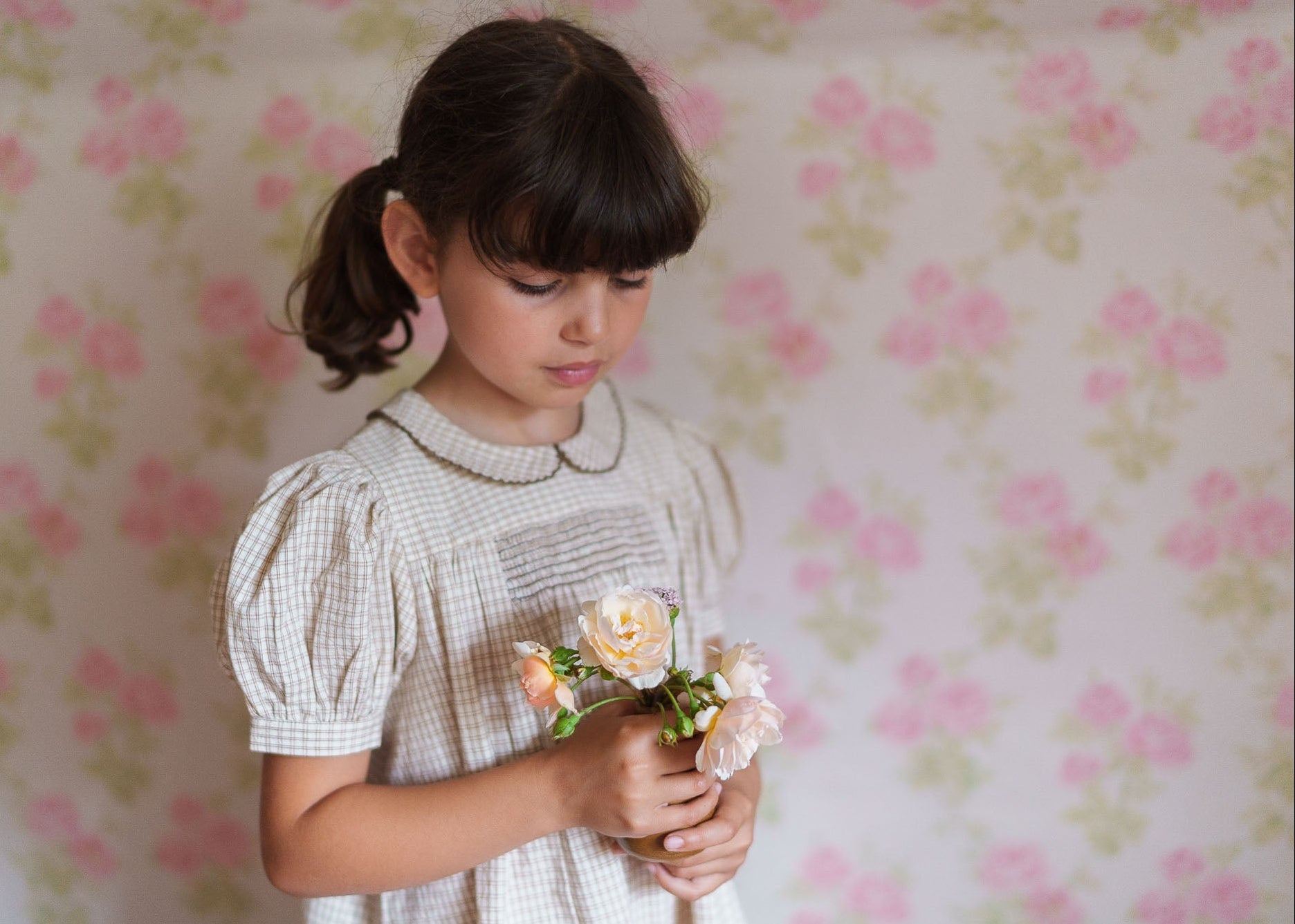 Young girl wearing a vintage style yellow checkered cotton blend dress holding delicate flowers against a soft floral wallpaper background in a natural, ethical children's clothing setting.
