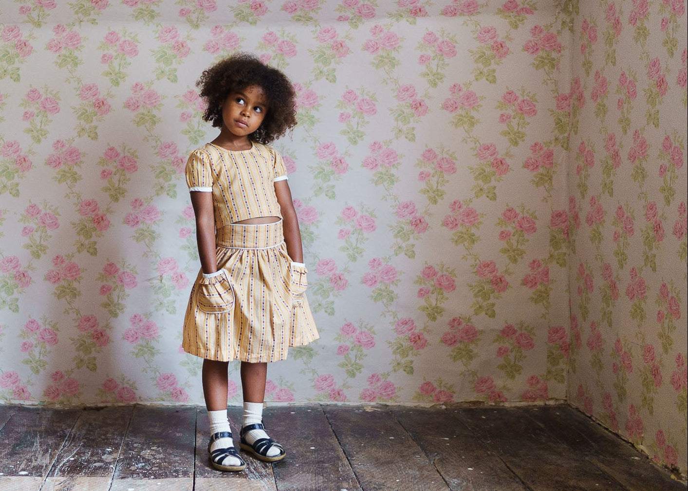 Young girl wearing a vintage style organic cotton floral top and skirt set, standing on rustic wooden floor against soft pink floral wallpaper, embodying natural and ethical children's clothing.