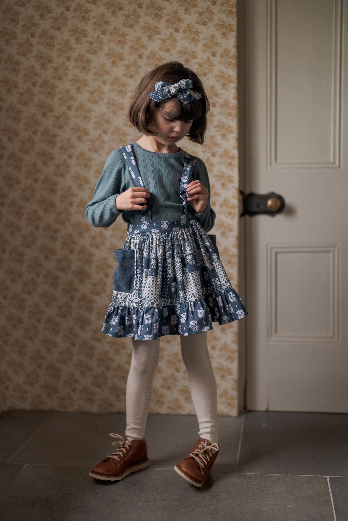 Young girl wearing a navy blue vintage style patchwork skirt with suspenders over a slate-colored cotton long-sleeve top, paired with cream tights and brown leather shoes in a classic interior.