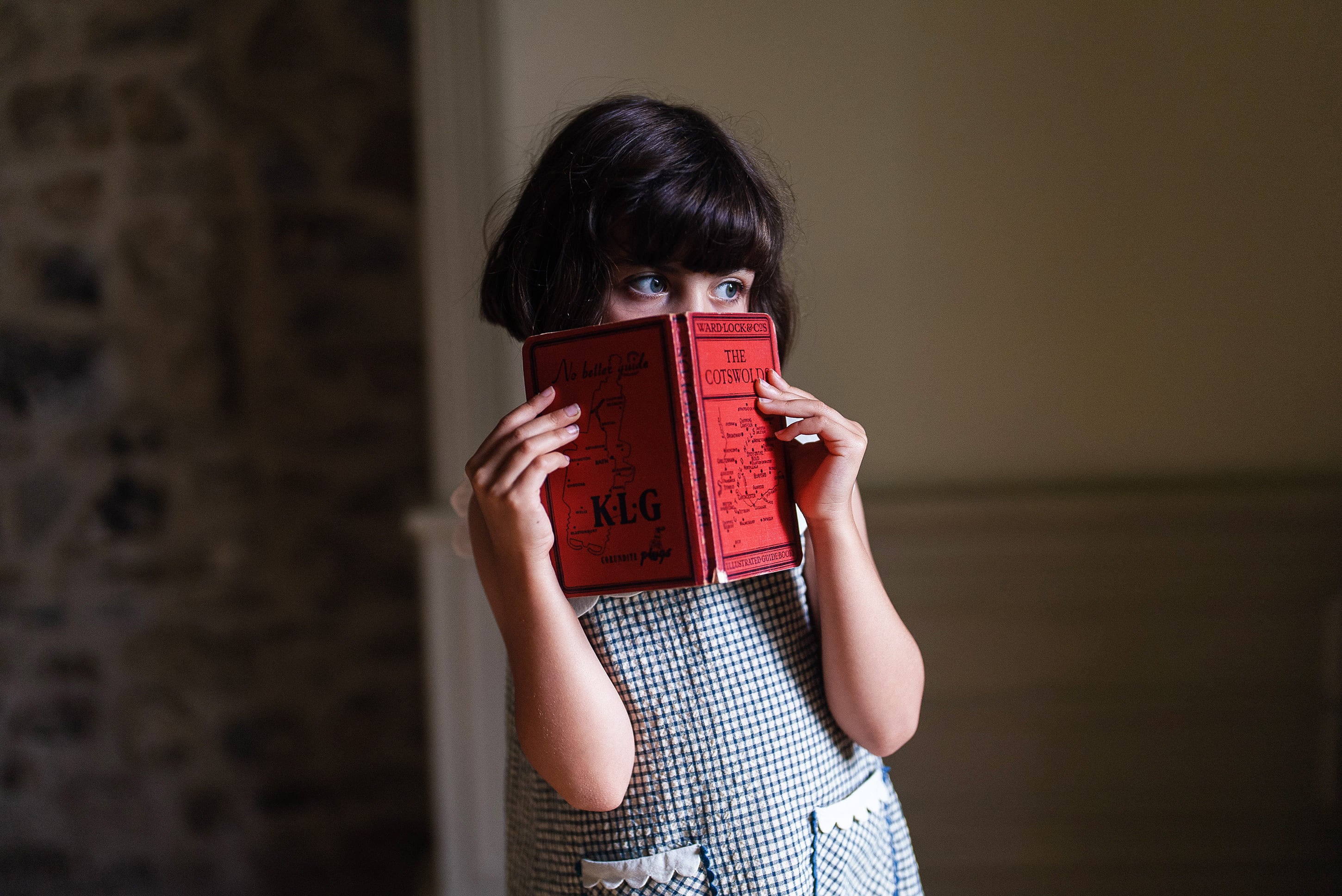 Young girl wearing a vintage style navy and white checkered cotton dress holding a small red book, with natural soft lighting creating a warm, cozy atmosphere.