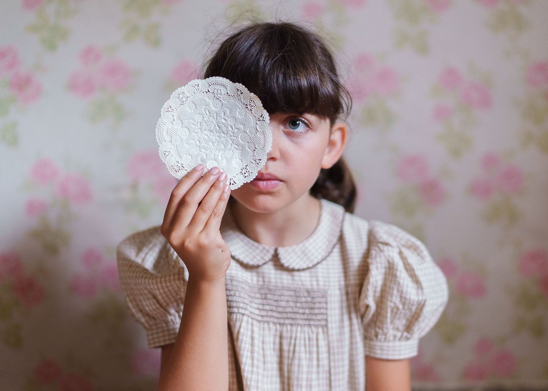 Young girl wearing a vintage-style checkered cotton dress in soft yellow tones, holding a delicate lace doily in front of her face against a floral wallpaper backdrop.